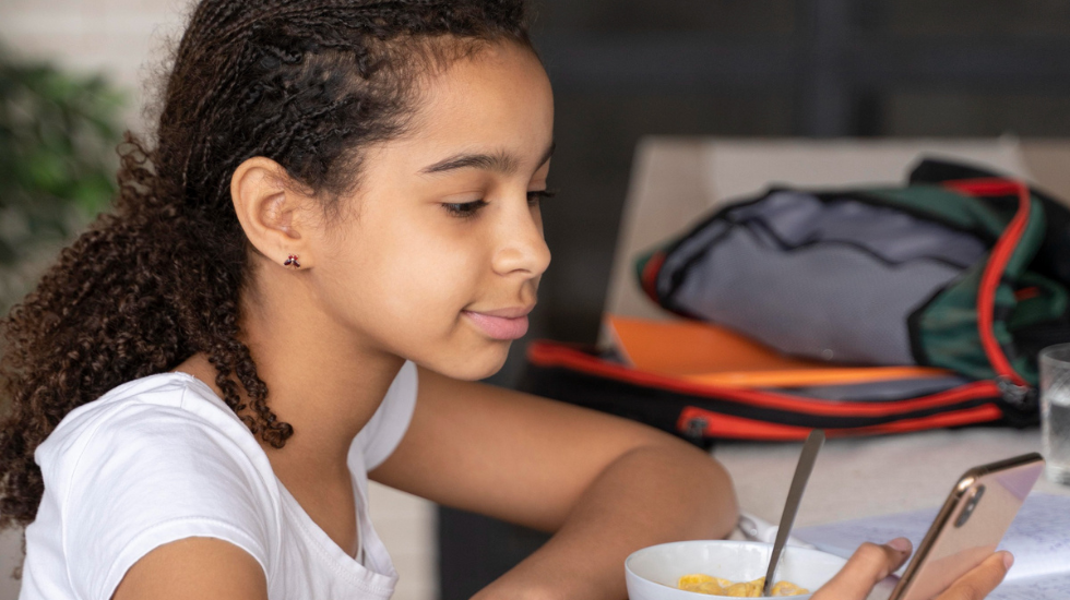 Menina se alimenta e usa celular na escola.